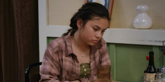 Still from The Moon and the Night. Mahina sits alone at a table in a restaurant.