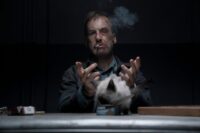 Hutch lets go of a cat to step forward and eat while he is handcuffed and smoking a cigarette.