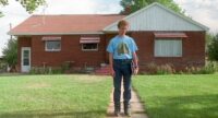Napoleon (Jon Heder) standing outside his house. He is wearing puffy boots and a blue shirt with horses on it tucked into his jeans. The lawn is freshly cut and the russet roof of the brick home is cast in sunlight.