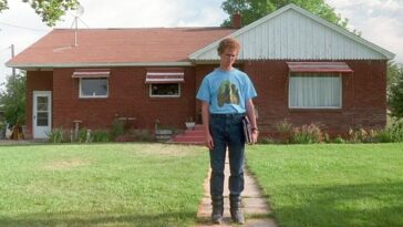 Napoleon (Jon Heder) standing outside his house. He is wearing puffy boots and a blue shirt with horses on it tucked into his jeans. The lawn is freshly cut and the russet roof of the brick home is cast in sunlight.