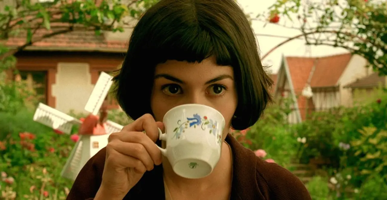 Amelie (Audrey Tautou), a white girl with dark eyes and a black bob hairstyle, holding a white teacup with a blue foral design up to her mouth. She is sitting outside in a garden, in front of a miniature windmill.