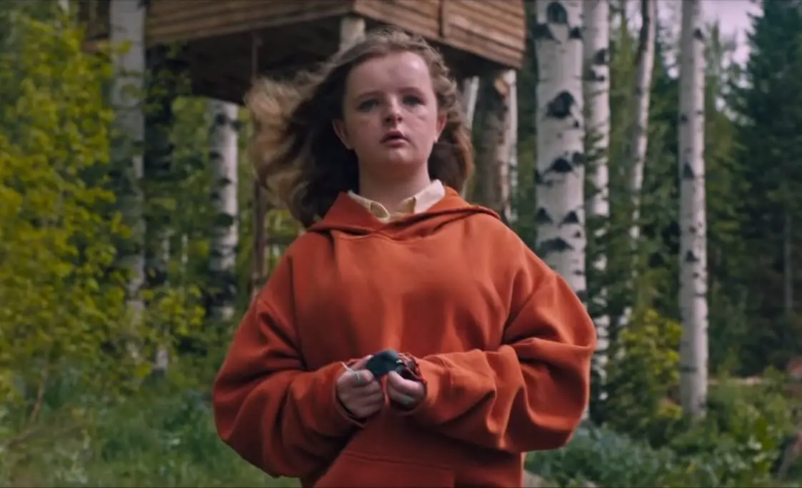 Charlie (Milly Shapiro), a white girl with long dirty blonde hair and blue eyes, standing in front of a tree house and a forest of birch trees. She is wearing an orange-ish red hoodie and is holding what appears to be a dead bird. She has two green strings tied around two different fingers.