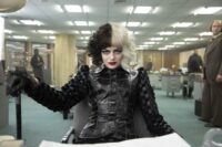 Cruella sits at a reporter's desk in an office.