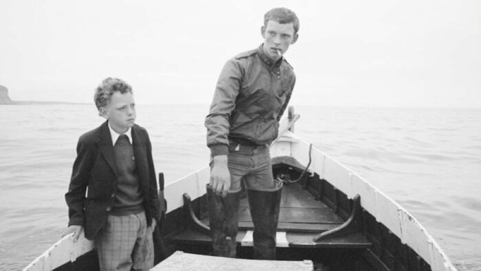 Still from Skinningrove depicting one of the photographs shown in the film. A young boy sits in a boat with an older boy. The young boy is wearing nice clothes and almost looks shell shocked. The older boy is smoking a cigarette.