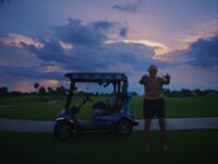 A shortless man stands next to a golf cart at dusk.