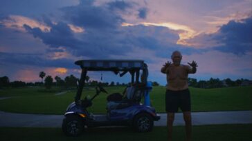 A shortless man stands next to a golf cart at dusk.