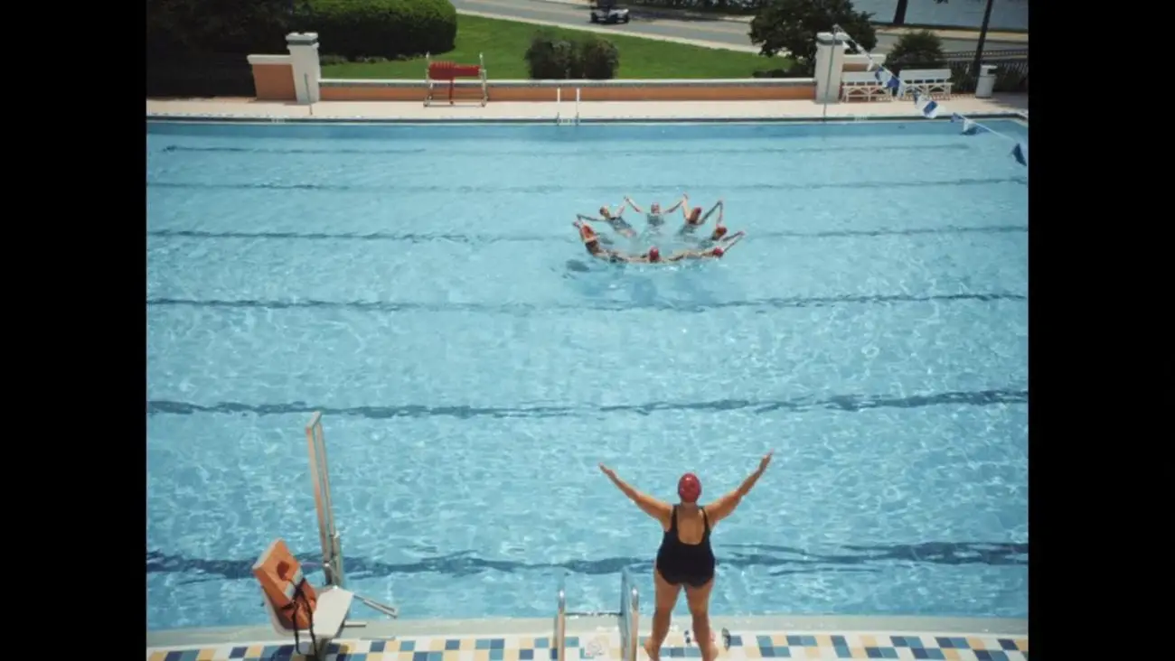 A woman directs a group of synchronized swimmers in a ppol.