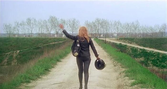 The blonde in black leather waves goodbye while walking forward down a gravel pathway