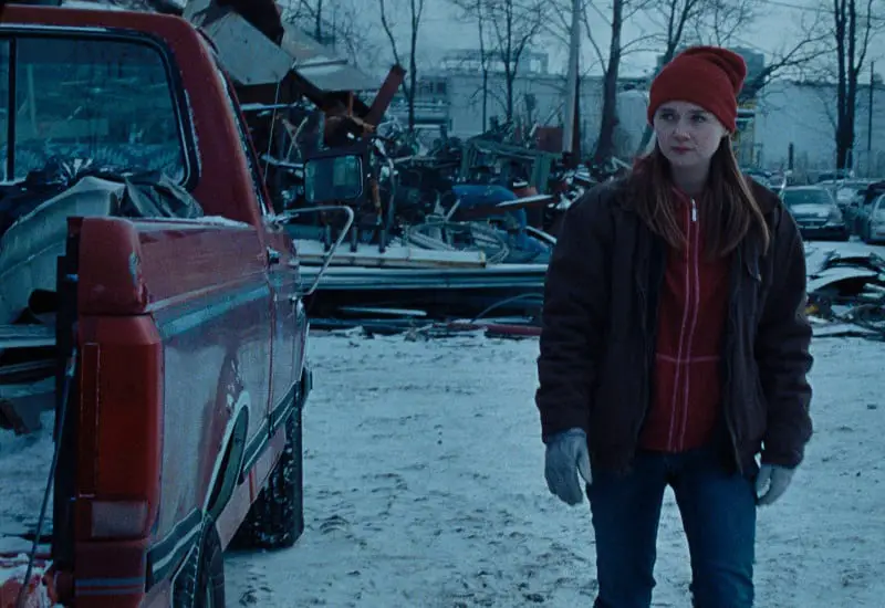 In Nicole Riegel's Holler, Ruth (Jessica Barden) stands next to a pickup truck in front of a metal scrapyard.