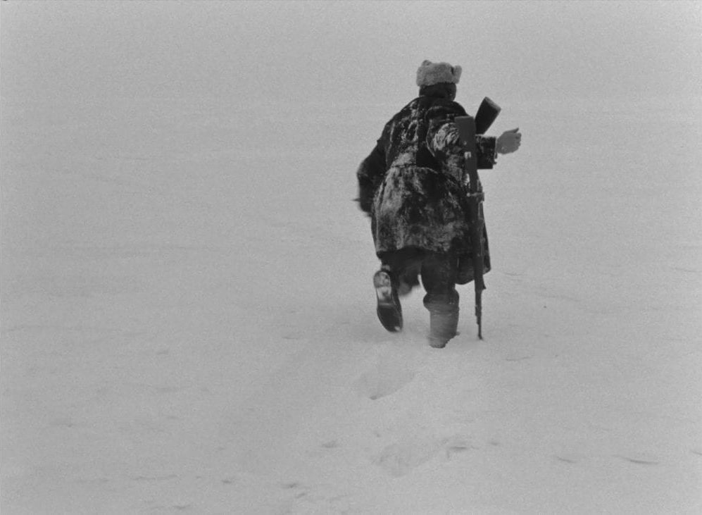 A man in a heavy coat with a rifle trudges through a deep barren snowdrift in this image from The Ascent..