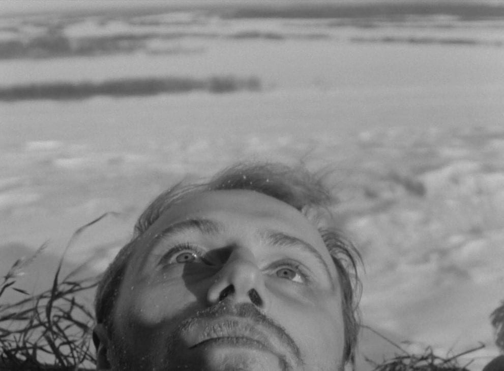 His head framed by strands of straw, the character Sotnikov gazes upward at the sky, a snowy field behind him in this image from The Ascent.