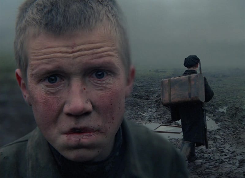 In this image from Come and See, the character Flyora, aged by his experience, stares in angusih at the camera while a young boy behind him carries a rifle and pack.