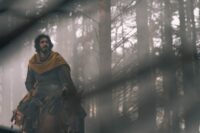 Gawain rides on horseback through a sunlight forest.