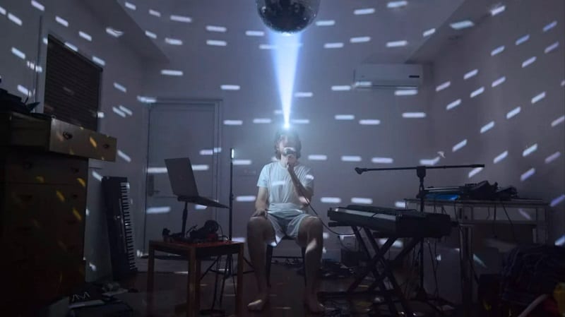 In this image from Inside, Bo Burnham is depicted alone in a room, a headlamp aimed at a mirrored ball creating a lightshow.