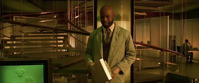 In this image from Gattaca, a geneticist played by Blair Underwood is depicted in a white laboratory coat in front of a spiral staircase in a modern office building.