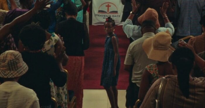 Screenshot from the Criterion Channel short film Lizard. Juwon stands in the aisle of a crowded church.