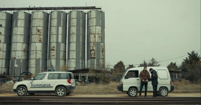 Screenshot from the Criterion Channel Short Sticker. A man stands outside of his car in an extreme wide shot with a police officer. They are in front of some industrial silos.