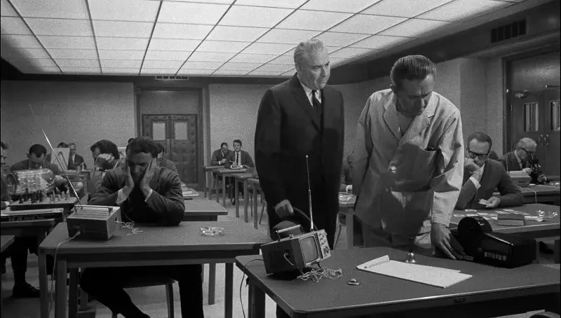 In this image from Seconds, Arthur Hamilton (John Randolph) is depicted in long shot in a room asking for help in a room where dozens of men sit quietly reading and watching portable televisions at their desks.