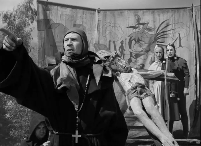 In this image from The Seventh Seal, a monk (played by Anders Ek) addresses an audience as Jof and MIa listen in costume from their stage alongside an effigy of Christ on the cross.