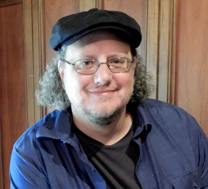  Steve Anderson wears a hat, glasses and has on a soft-toned collared shirt with the buttons open with a black t-shirt underneath. There is wood paneling behind him