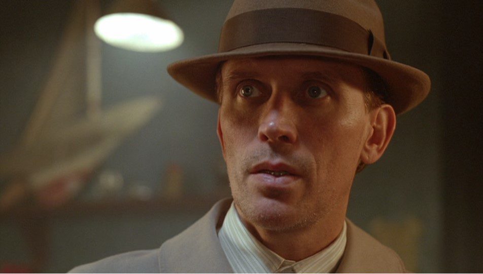Bill Lee (Peter Weller) wears a khaki jacket, hat, and suit and has a shocked expression on his face.