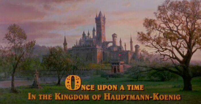A castle stands with a pastel sky behind it as words on the bottom of the screen read "Once upon a time in the kingdom of Hauptmann-Koenig"
