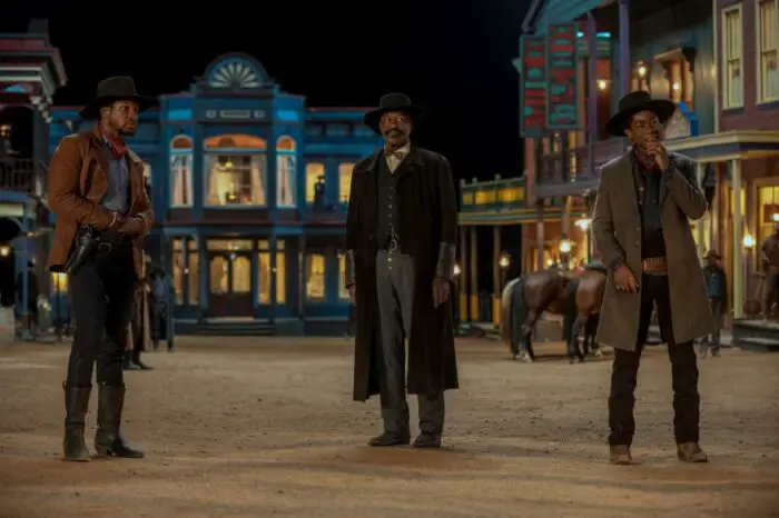 Three men in western wear stand and watch in a well-lit street.