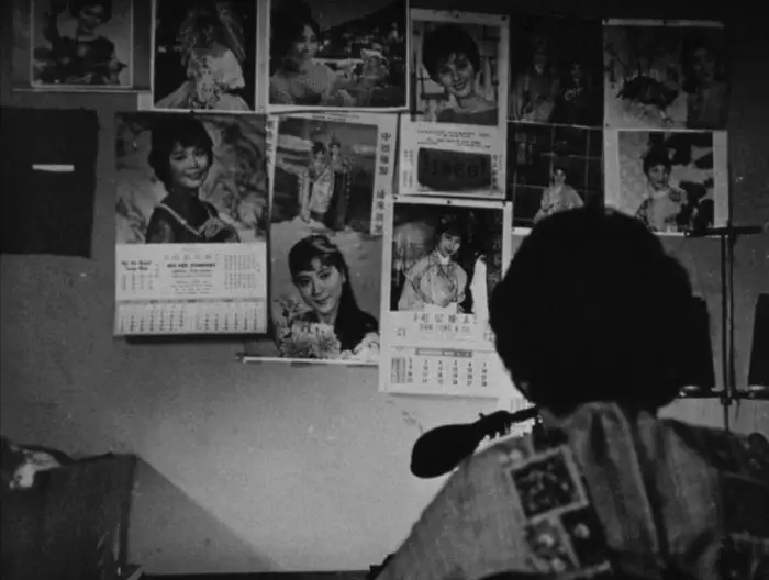 Screenshot from Sewing Woman, one of the short films added to Criterion Channel in October. A woman sits in front of a wall of calendars and pictures. The shot is black and white.