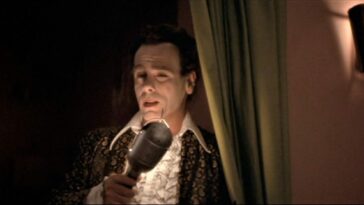 A man wearing a white frilled shirt with a large collar and a gold and black blazer holds a single light bulb to his face like a microphone (Dean Stockwell in Blue Velvet)