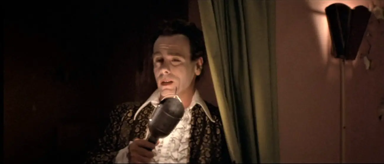 A man wearing a white frilled shirt with a large collar and a gold and black blazer holds a single light bulb to his face like a microphone (Dean Stockwell in Blue Velvet)