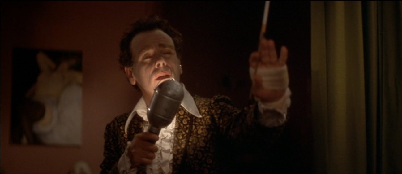 A man wearing a frilled shirt, patterned jacket holds a light bulb like a microphone in his right hand. His left hand is bandaged and also holds a cigarette holder. (Dean Stockwell mimes in Blue Velvet)