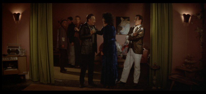 Several people stand in a dark room framed by green curtains and two lamps affixed to pale pink walls; A man in a black leather jacket (Dennis Hopper) places his hand on a woman wearing a dark blue robe (Isabella Rossellini). A man stands beside them holding a lightbulb (Dean Stockwell).