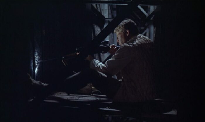 A man sits in a dark room aiming a rifle which is pointed through a wall.