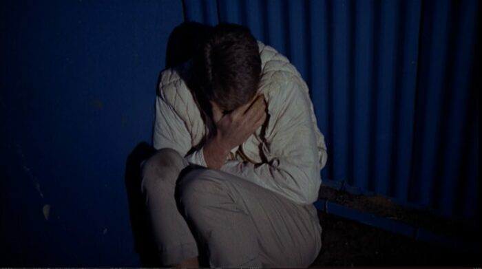 A man wearing dirty slacks and a beige jacket sits curled up in a ball surrounded by a corrigated metal wall.