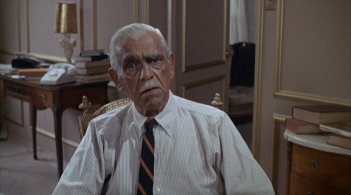 An old man with white hair and a white mustache wearing a shirt and tie sits in a hotel room and stares at the viewer.