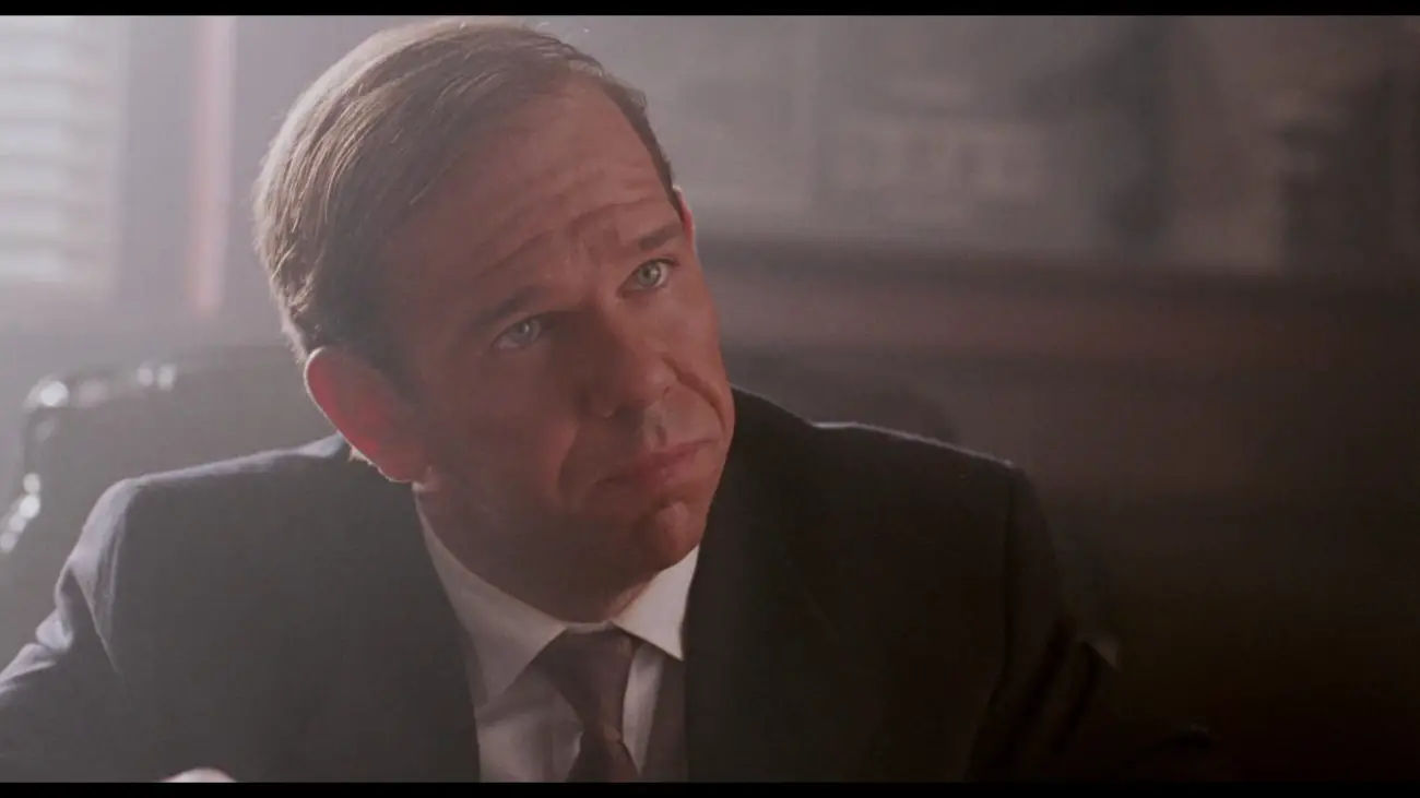 Image from Deep Cover: DEA Officer Carver (Charles Martin Smith) is depicted in close-up dressed in a suit and tie in an office with venetian blinds.