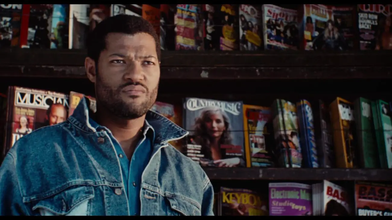 Image from Deep Cover: Stevens/Hull (Fishburne) stands in front of a magazine stand, wearing a denim jacket.