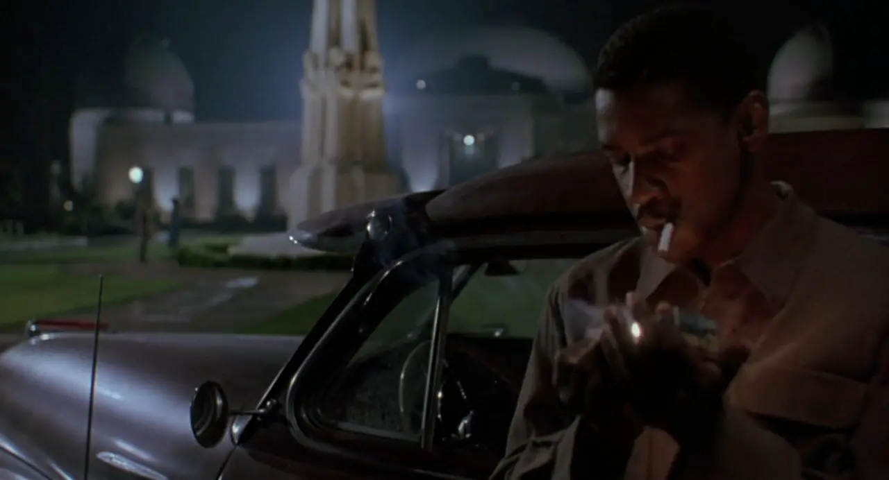 Image from Devil in a Blue Dress: Easy Rawlins (Denzel Washington) lights a cigarette outside his parked car at the Griffith Observatory at night, the characters of Daphne Monet (Jennifer Beals) and Todd Carter (Terry Kinney) in the background.