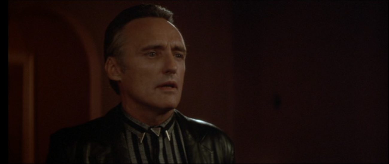 A man wearing a black leather jacket, striped shirt, and triangular metallic bolo tie stands in a darkened room and stares longingly into the distance. (Dennis Hopper as Frank Booth in Blue Velvet)