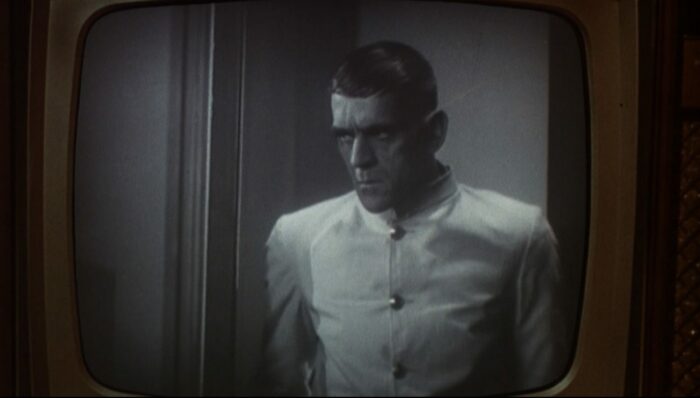 A menacing man wearing a jacketed with large buttons looms in a doorway; the image is framed by the exterior of a television screen, and the film is in black and white (Boris Karloff in The Criminal Code)