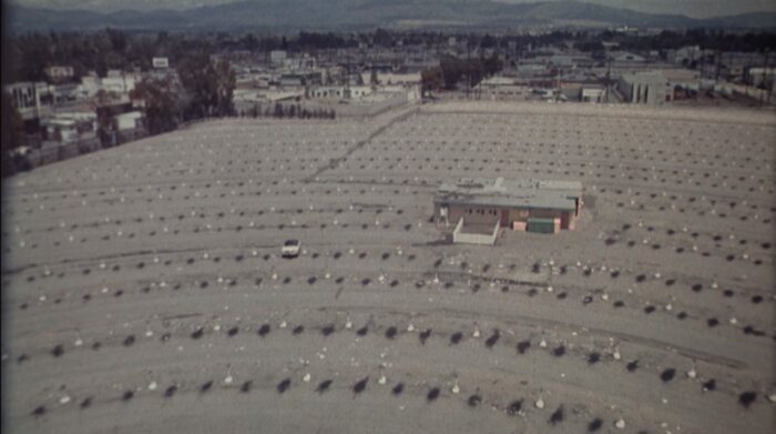 An aerial view of a large, vacant drive-in movie theater parking lot during the daytime; a single white car sits in the lot.