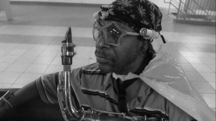 Screenshot from The Dreamer, one of the short films added to Criterion Channel in November. A man wearing a hat, flashlight, and trashbag cape holds a saxophone as he stares off camera.