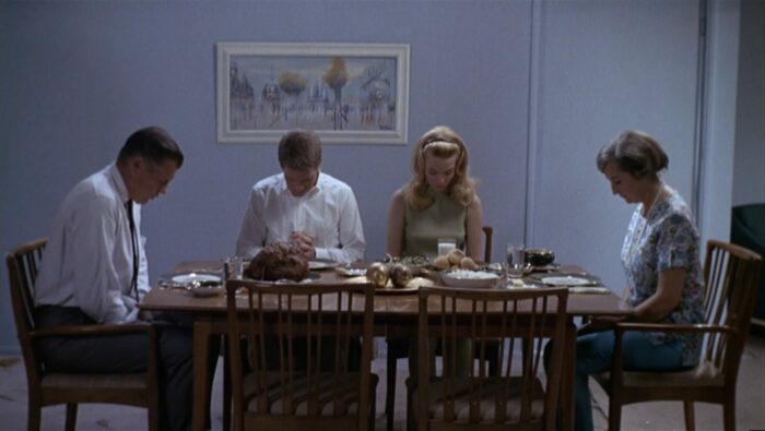 Four adults sit with their heads bowed at a wooden table, which is covered with food (The Thompson family has dinner in the film Targets)