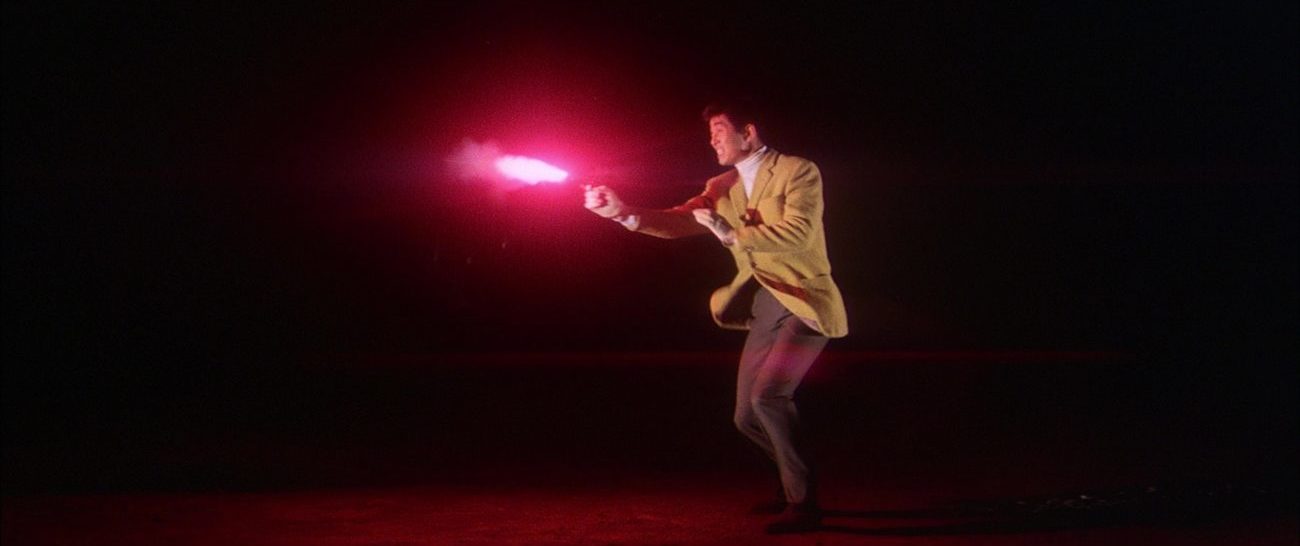 Image from Tokyo Drifter: Depicted in long shot against a black background, Tetsu fires his pistol with a bright crimson flash.