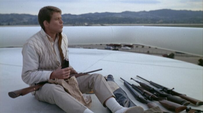 A young white man wearing khaki clothing sits atop a white oil tanker next to several rifles; he is holding a bottle of soda, looking off into the distance (Tim O'Kelly in Peter Bogdanovich's Targets).