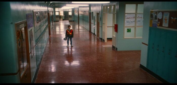 Greg in his safety patrol uniform standing alone in the empty school hallway. The floor is a reddish brown and the walls are turquouse.