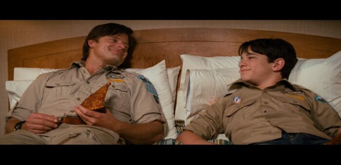 Frank (right) and Greg (left), both wearing beige wilderness explorer uniforms, laying on a motel bed eating pizza. They are smiling at each other.