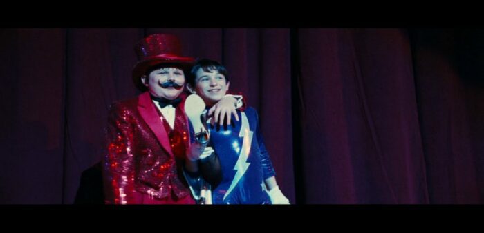 Rowley Jefferson (Robert Capron, left) wearing a red sequin suit and top hat with his arm around Greg (right), who is wearing a dark blue leotard with a white thunderbolt design across the front. Greg is holding up a prop high-heeled and fish-netted leg. The pair are on stage at their town's talent show, standing in front of a dark berry red curtain.