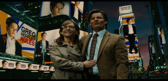 Susan Heffley (Rachael Harris), a white woman with long brown hair wearing glasses and a beige trench coat with Frank Heffley (Steve Zahn), a white man with dark brown hair wearing a brown suit and tie with a light blue button-up shirt standing outside in a fantasy version of Times Square. They are surrounded by images of their son, Greg. The sky is a mix of blue and lavender.