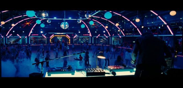 A roller rink lit by deep blue light. The camera is positioned behind the DJ's table looking out over the busy rink. There are various glowing balls of light hanging from the ceiling.